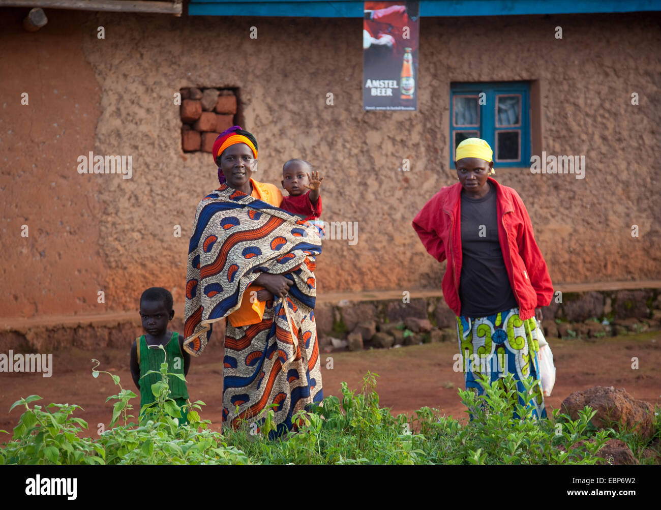I passanti in piedi di fronte ad un fango di umile casa di mattoni con una piccola comunità di birra poster pubblicitario sospesi dal tetto, Burundi, vicino al Parco Nazionale de la Ruvubu, Cankuzo Foto Stock