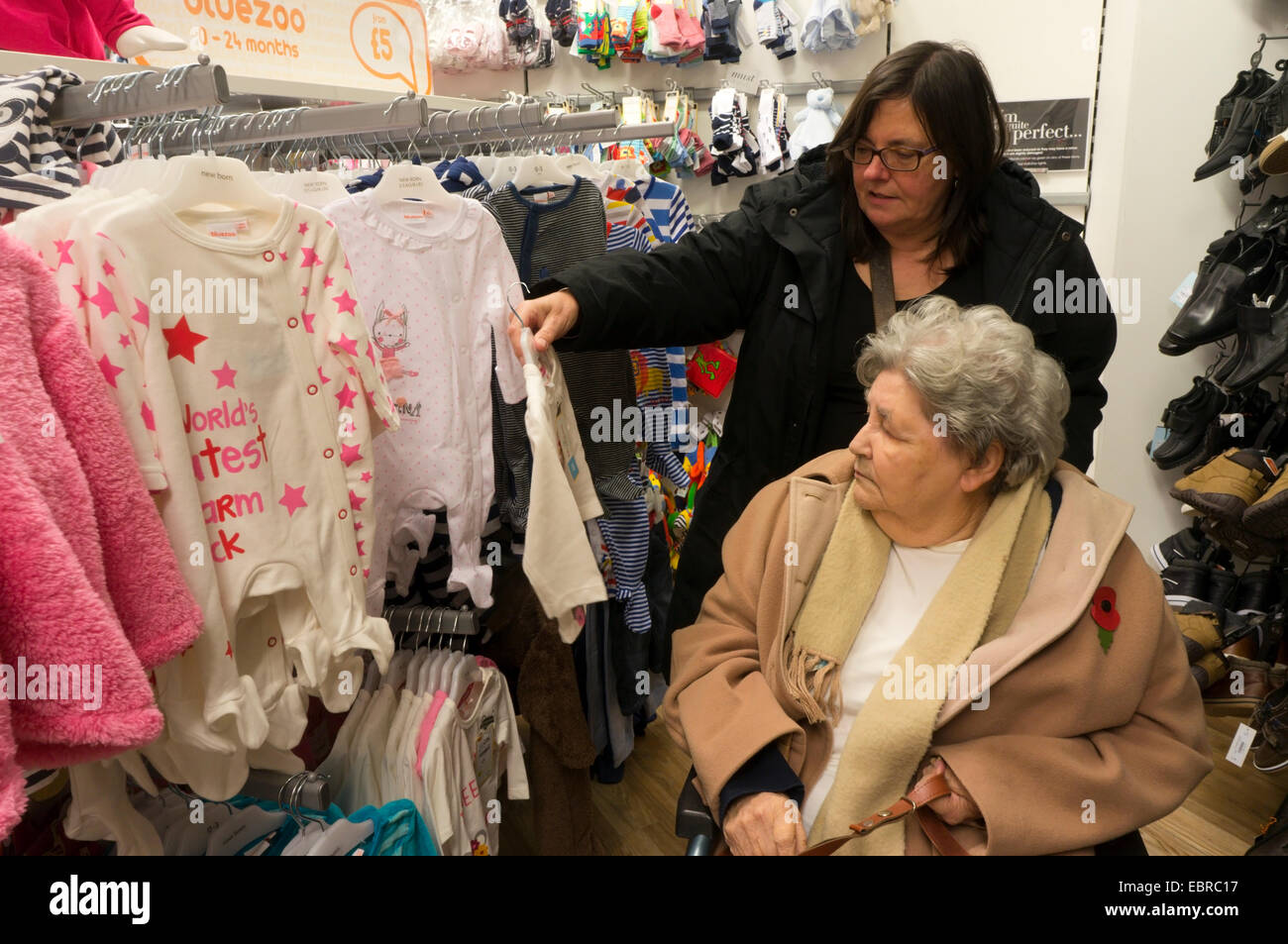Signora anziana in carrozzina e il suo accompagnatore o assistente shopping per vestiti del bambino in un grande magazzino. Foto Stock
