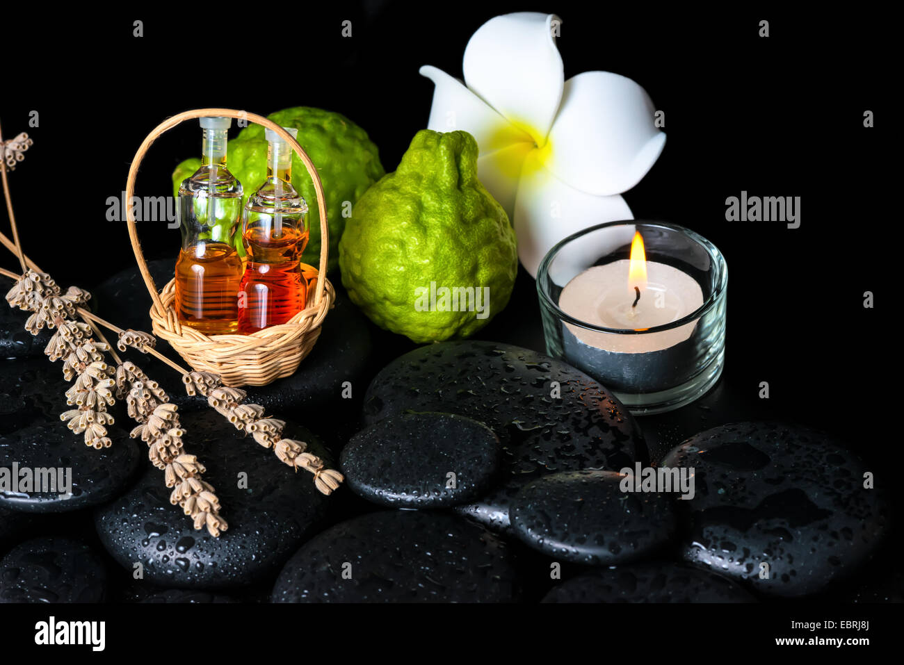 Centro termale aromatico nozione di bottiglie di olio essenziale di bergamotto, frutti, plumeria fiore, candela e lavanda essiccata su pietre zen Foto Stock