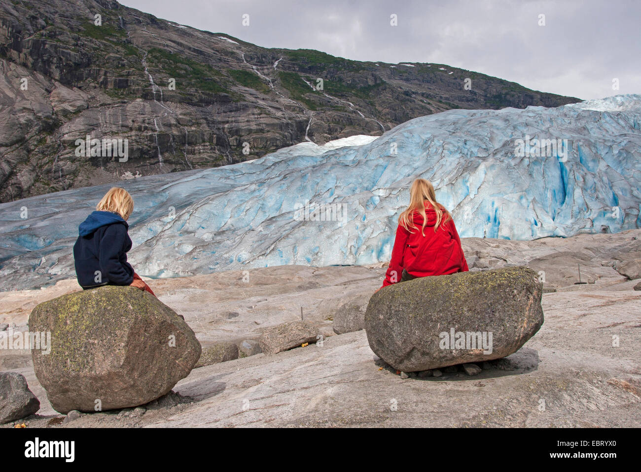 Due bambini seduti sulle rocce di fronte del ghiacciaio Nigardsbreen, Norvegia, Jostedalsbreen Parco Nazionale Foto Stock
