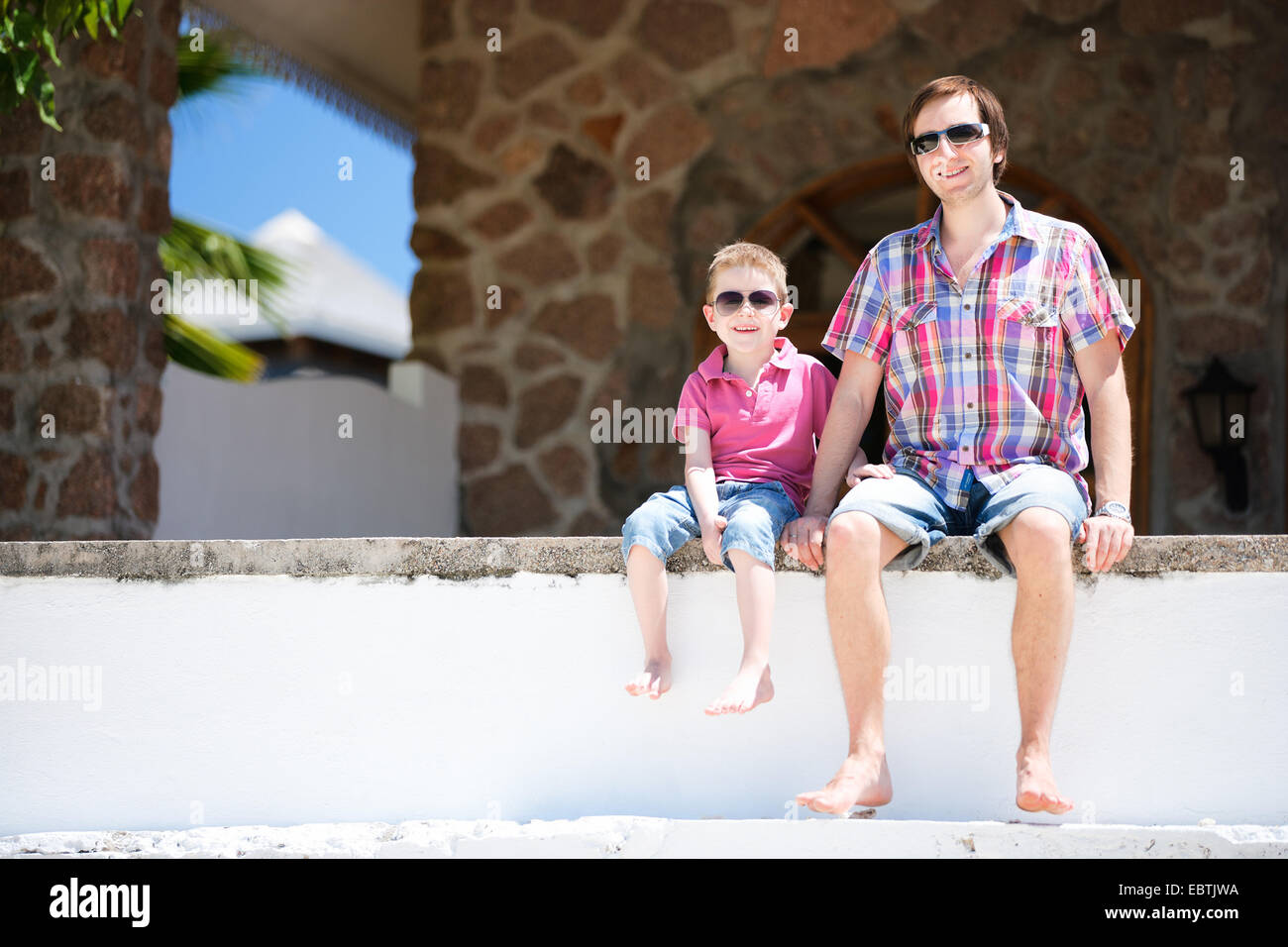 Padre e figlio seduti su una parete e guardando alla fotocamera Foto Stock