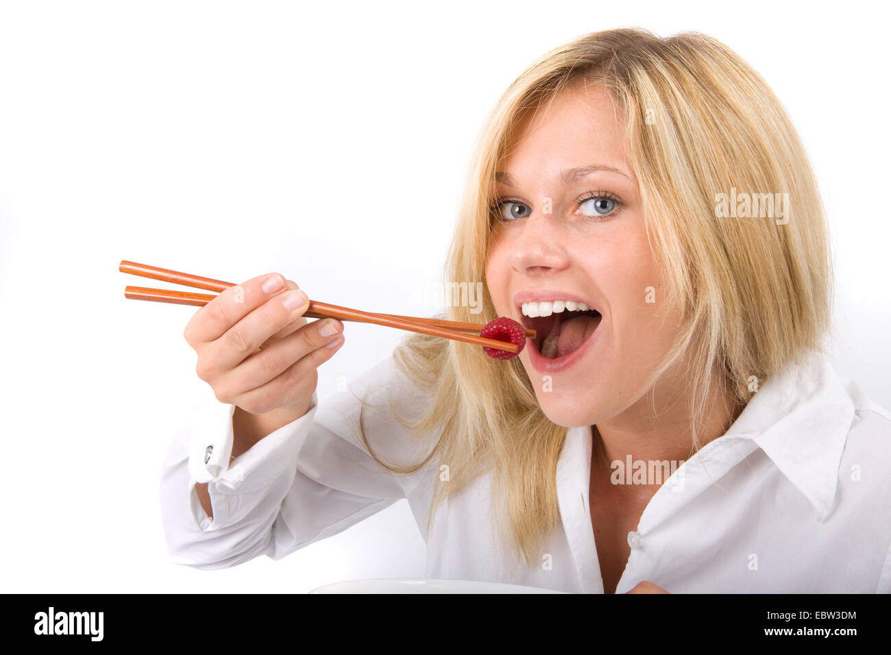 Giovane donna bionda sorridente mentre mangia un solo lampone da una piastra con bacchette Foto Stock