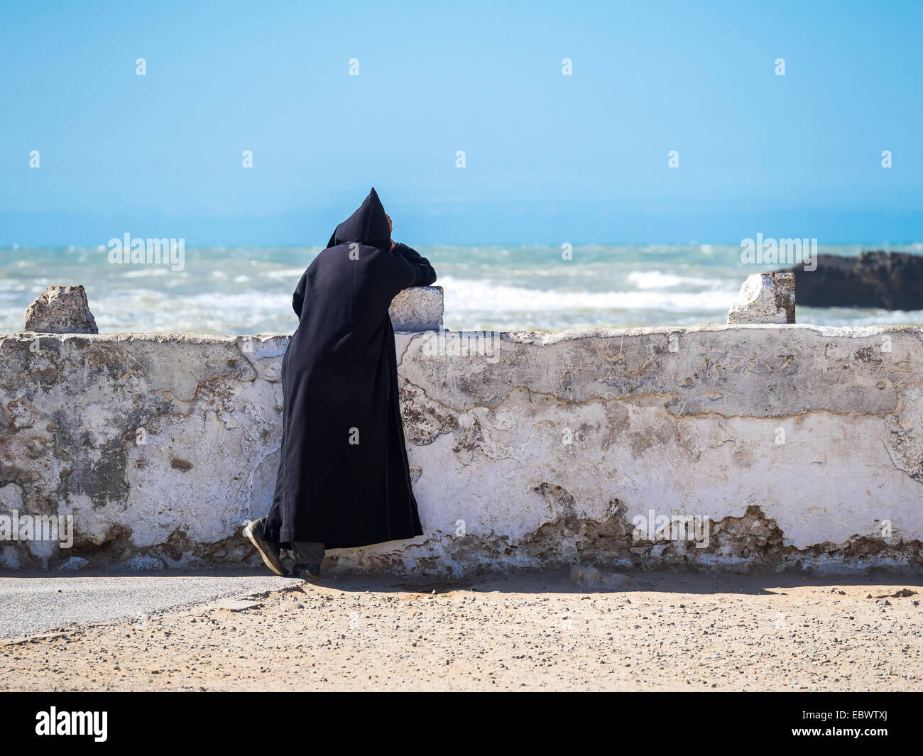 Un riff con djellaba nera appoggiata contro la parete del porto, place moulay Hassan, Essaouira, Marocco Foto Stock