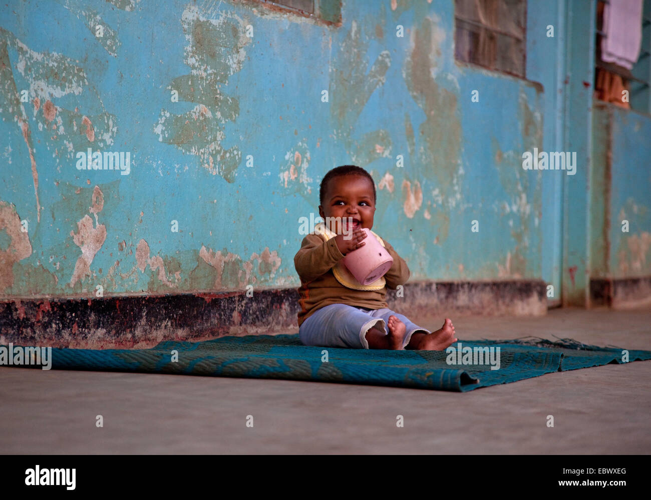 Baby sitter su un tappeto di fronte al muro squallido di un orfanotrofio aspirare ad un vaso in materia plastica, Burundi Bujumbura Marie, Bujumbura Foto Stock