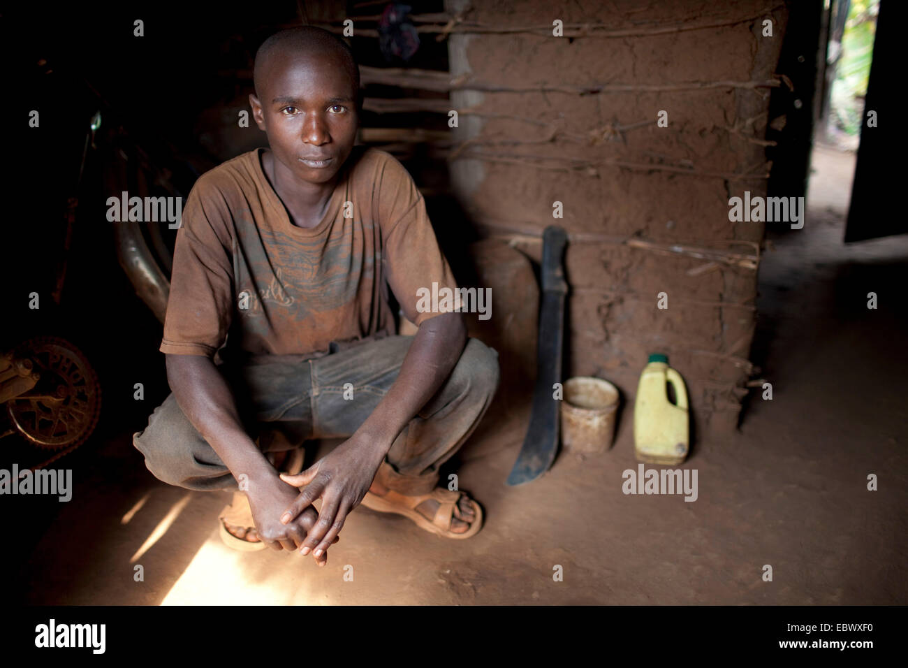 Giovane uomo squatting in ingresso della sua umile casa di fango con un machete accanto a lui, Burundi, Karuzi, Buhiga Foto Stock