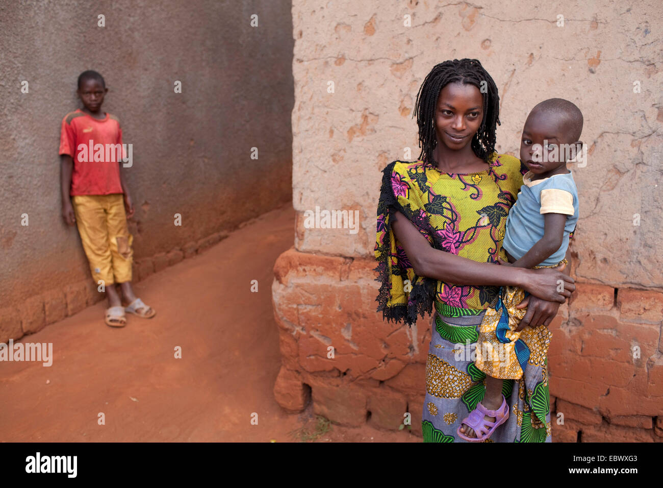 Giovane madre con piccolo figlio sui suoi bracci è in piedi in un vicolo di umile indossando abiti tradizionali, Burundi, Karuzi, Buhiga Foto Stock