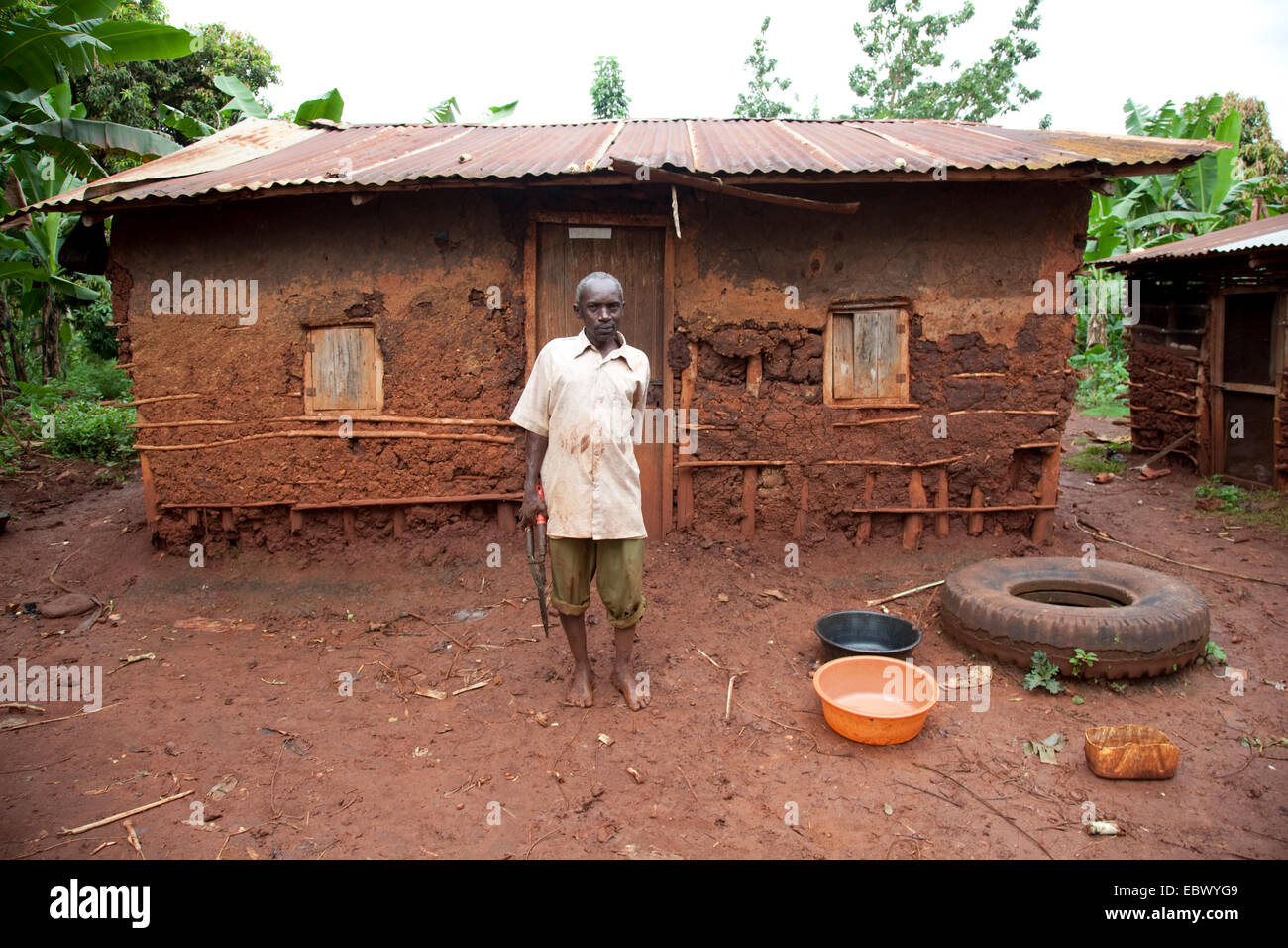 Uomo anziano è in piedi sulla parte anteriore della sua semplice casa di fango con un ferro corrugato tetto, Uganda, Jinja Foto Stock