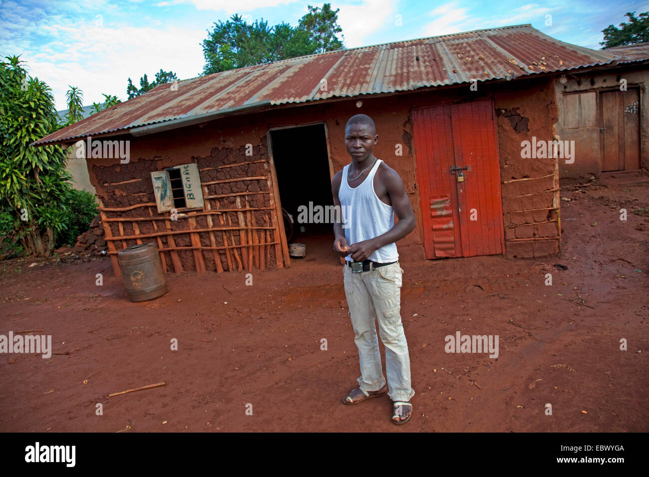 Giovane uomo è in piedi sulla parte anteriore della sua semplice casa di fango con un ferro corrugato tetto, Uganda, Jinja Foto Stock