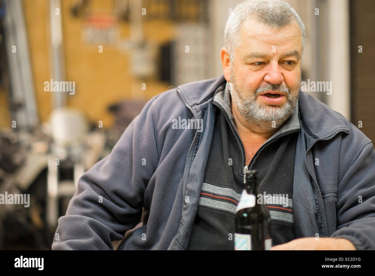 Un vecchio uomo caucasico tra 60 e 70 anni seduto nel suo negozio con bottiglia di birra a parlare con qualcuno. Foto Stock