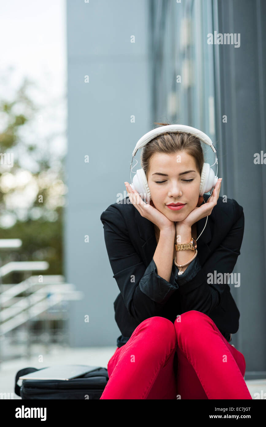 Giovane donna con gli occhi chiusi seduta sulle scale ascoltando musica con le cuffie Foto Stock