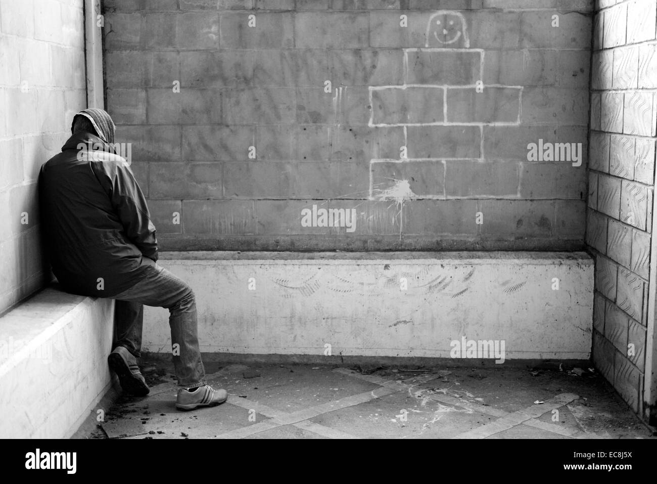 Uomo senza tetto rivolto lontano dalla telecamera ed il ricovero in un parcheggio sotterraneo, con graffiti sul muro. Monocromatico Foto Stock