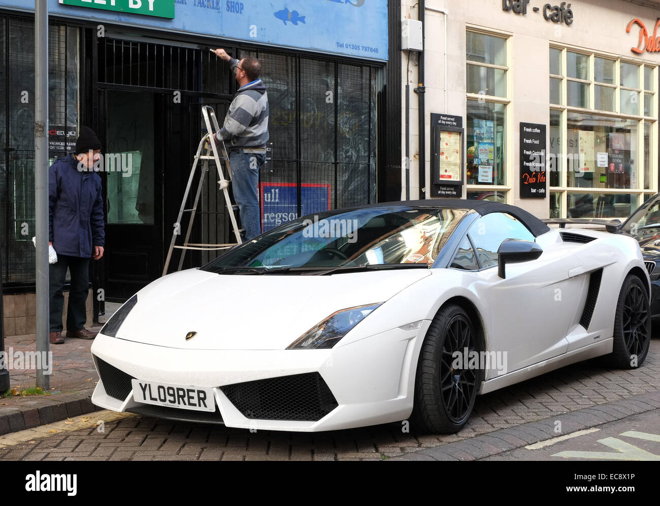 Il vecchio uomo e un uomo di verniciatura di un negozio di fronte a fianco di un esotico auto, una Lamborghini, Weymouth Town Center. 10 Dicembre 2014 Foto Stock