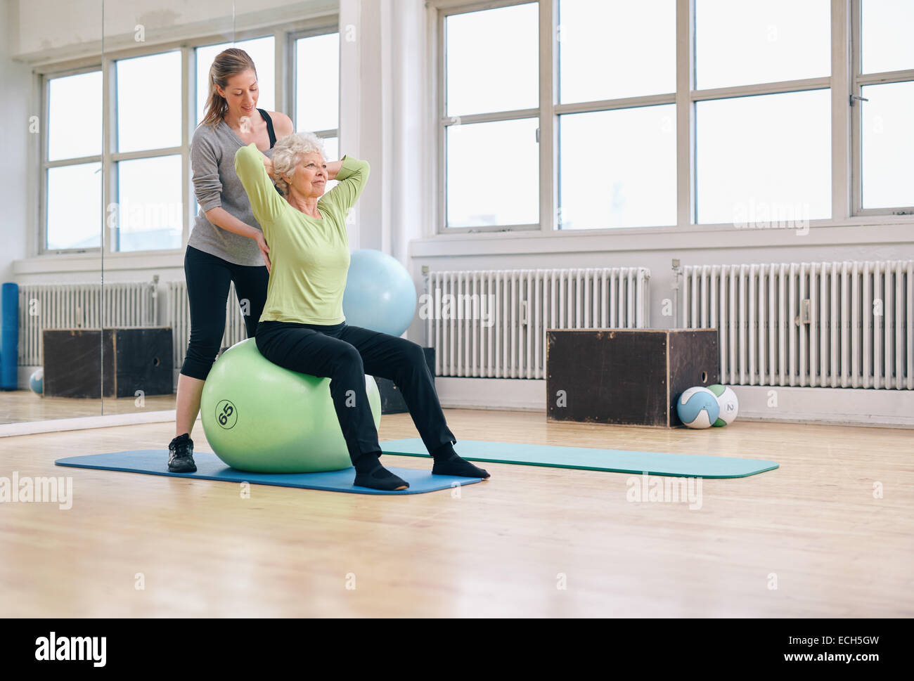 Senior donna seduta su una sfera di pilates esercizio al club della salute essendo coadiuvato dal suo personal trainer. Fisioterapista Foto Stock