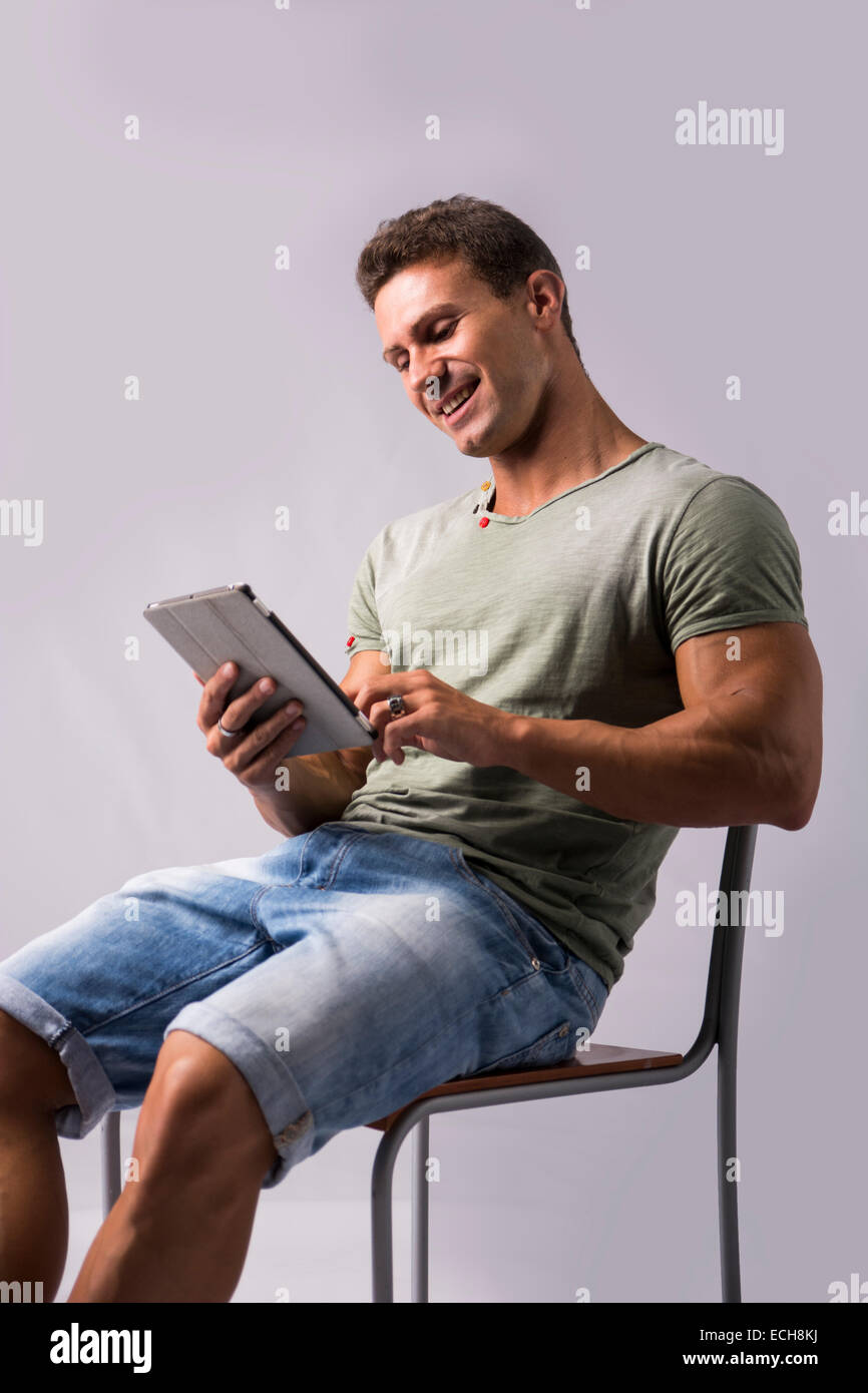 Muscolare di giovane uomo seduto su una sedia mentre la lettura da e-book dispositivo con felice espressione, isolato su bianco Foto Stock