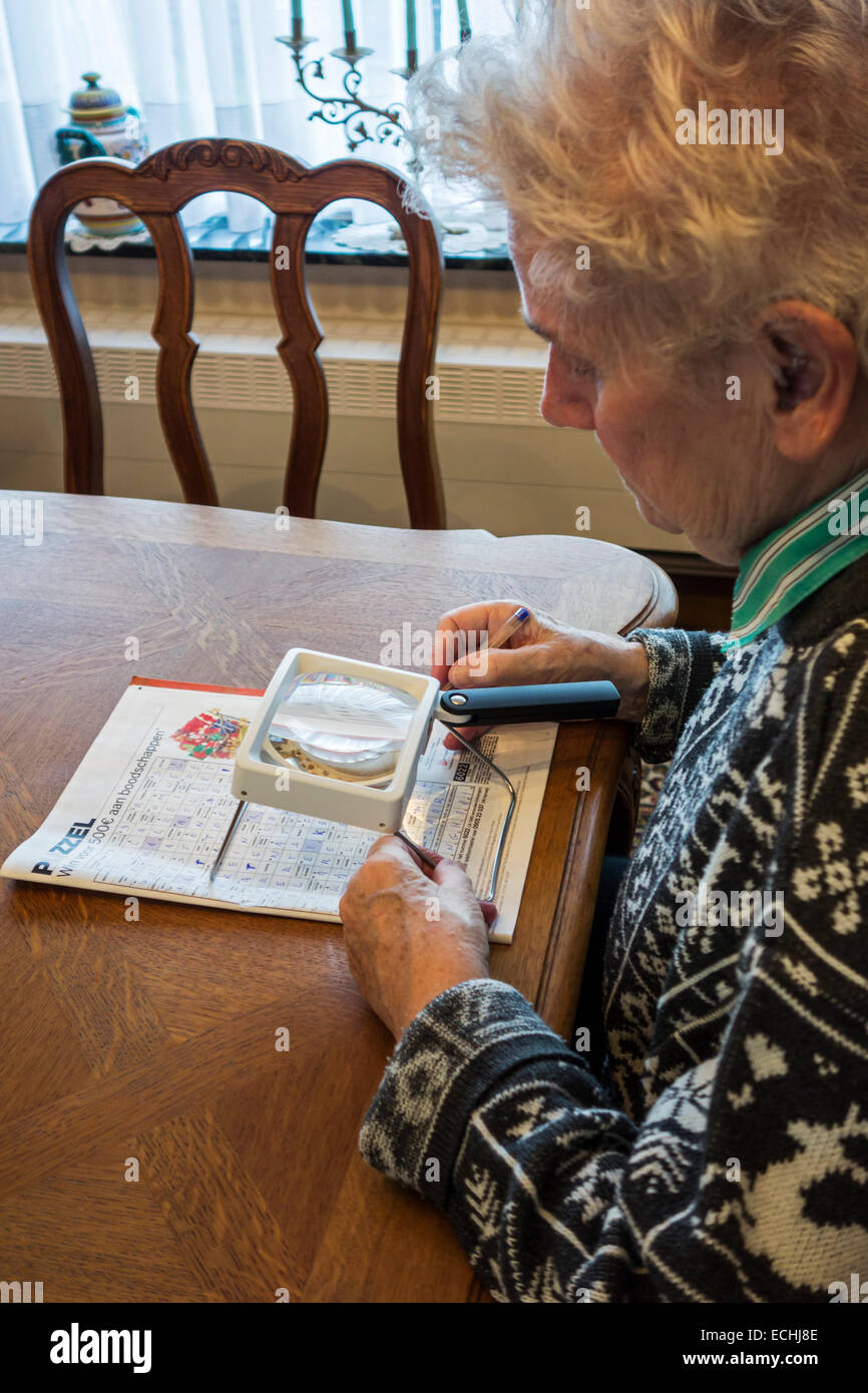 Donna anziana che soffre di presbiopia / miopia / miopia guardando cruciverba attraverso la lente di ingrandimento Foto Stock