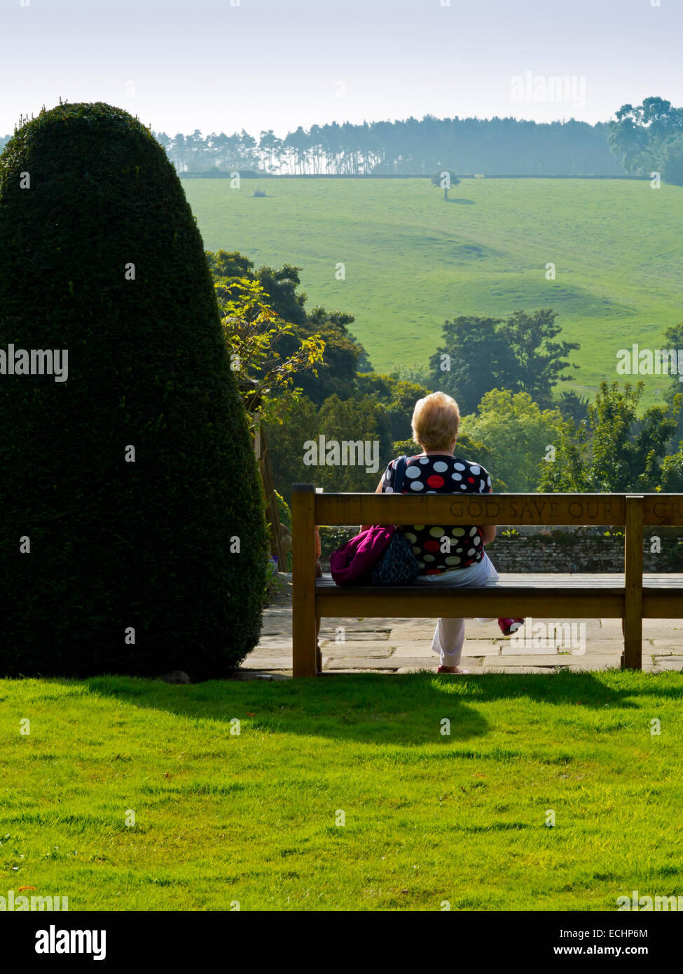 Donna anziana in un giardino seduta su una panchina rilassante e gode di vista sul paesaggio rurale accanto alla struttura topiaria da Foto Stock