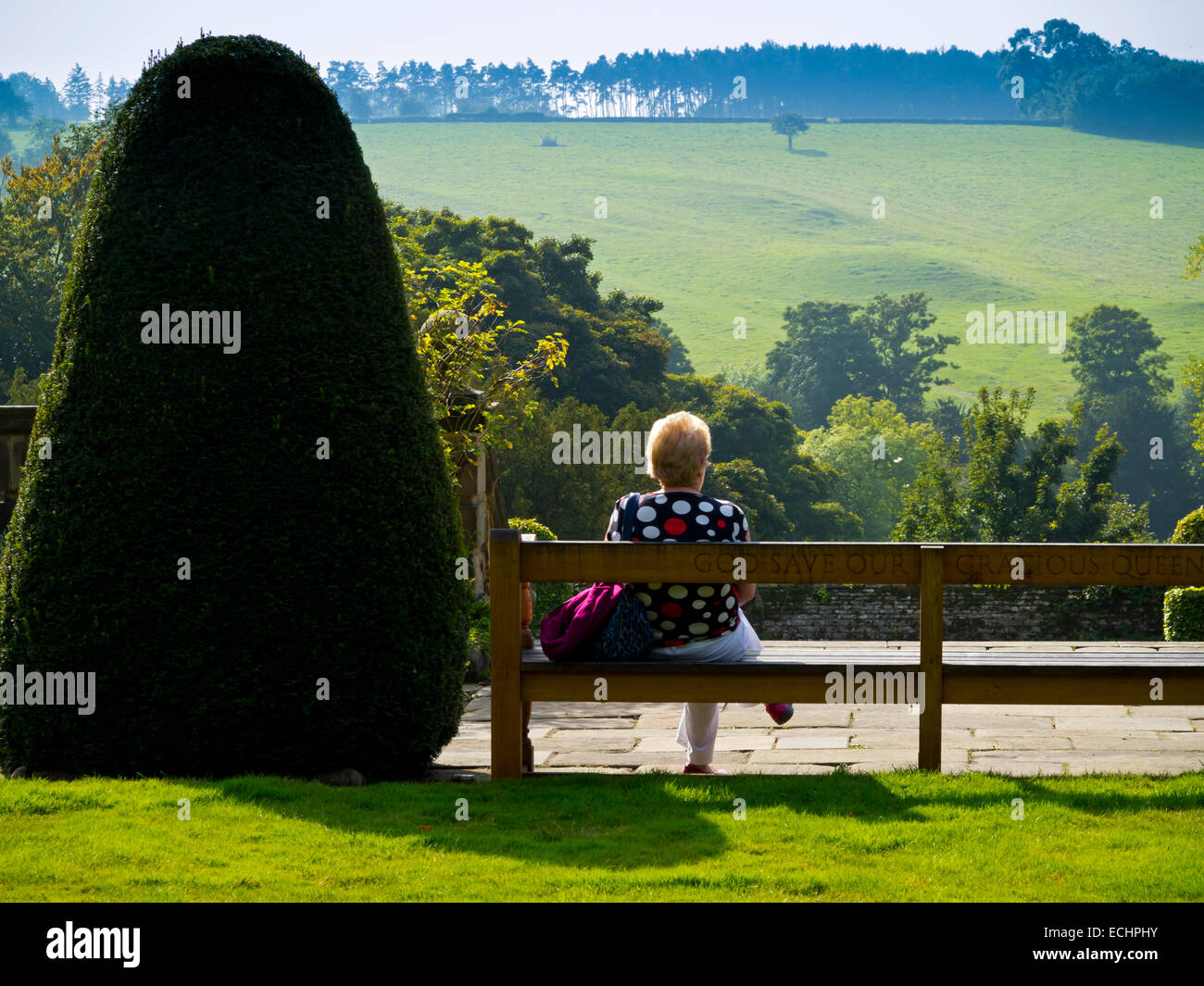 Donna anziana in un giardino seduta su una panchina rilassante e gode di vista sul paesaggio rurale accanto alla struttura topiaria da Foto Stock