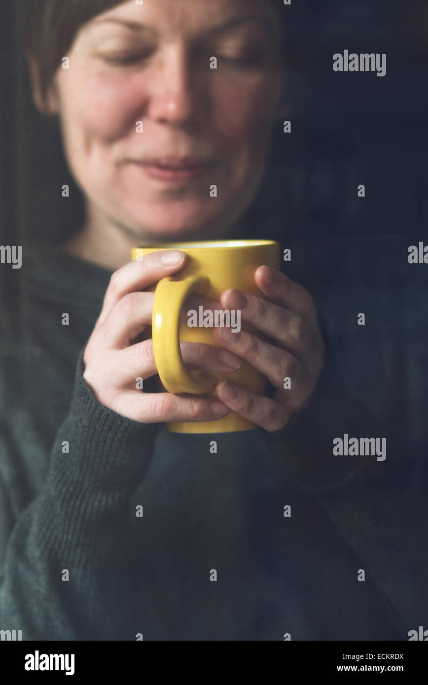 Bella adulto caucasica donna sorridente bere e godere di tazza di caffè nella stanza buia. Messa a fuoco selettiva su mani e la tazza con il Foto Stock