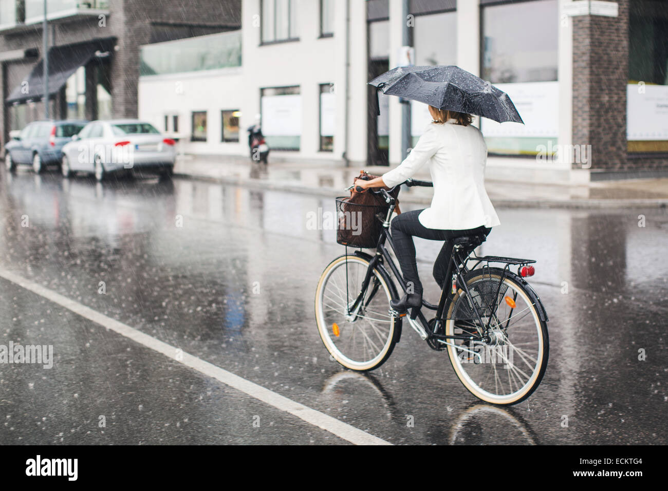 Lunghezza piena vista posteriore di imprenditrice Bicicletta Equitazione su bagnato via della città durante la stagione delle piogge Foto Stock