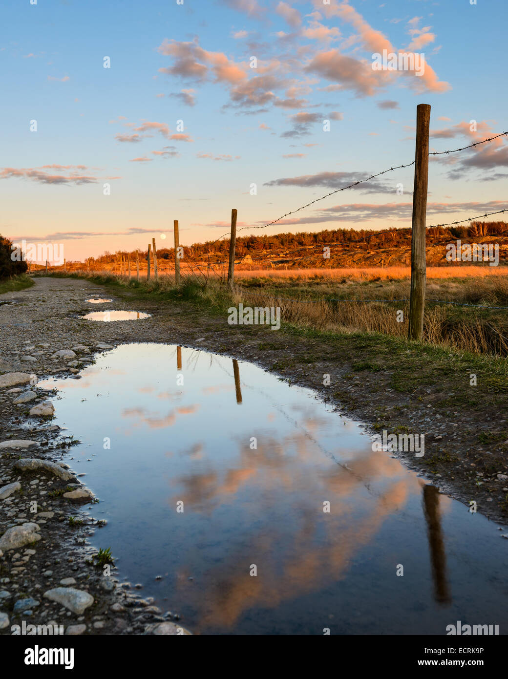 Nuvole e un agricoltore e la recinzione si riflette nelle pozzanghere sulla strada al tramonto Foto Stock