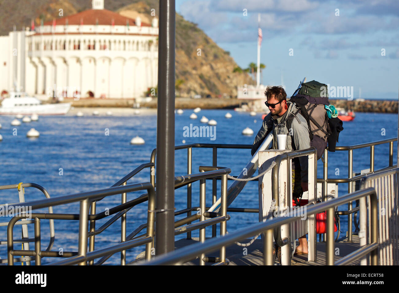 Giovane uomo porta una tavola da surf e Zaino grande sul Catalina traghetto espresso in Avalon, Isola Catalina, California.Lo storico Casinò dietro. Foto Stock