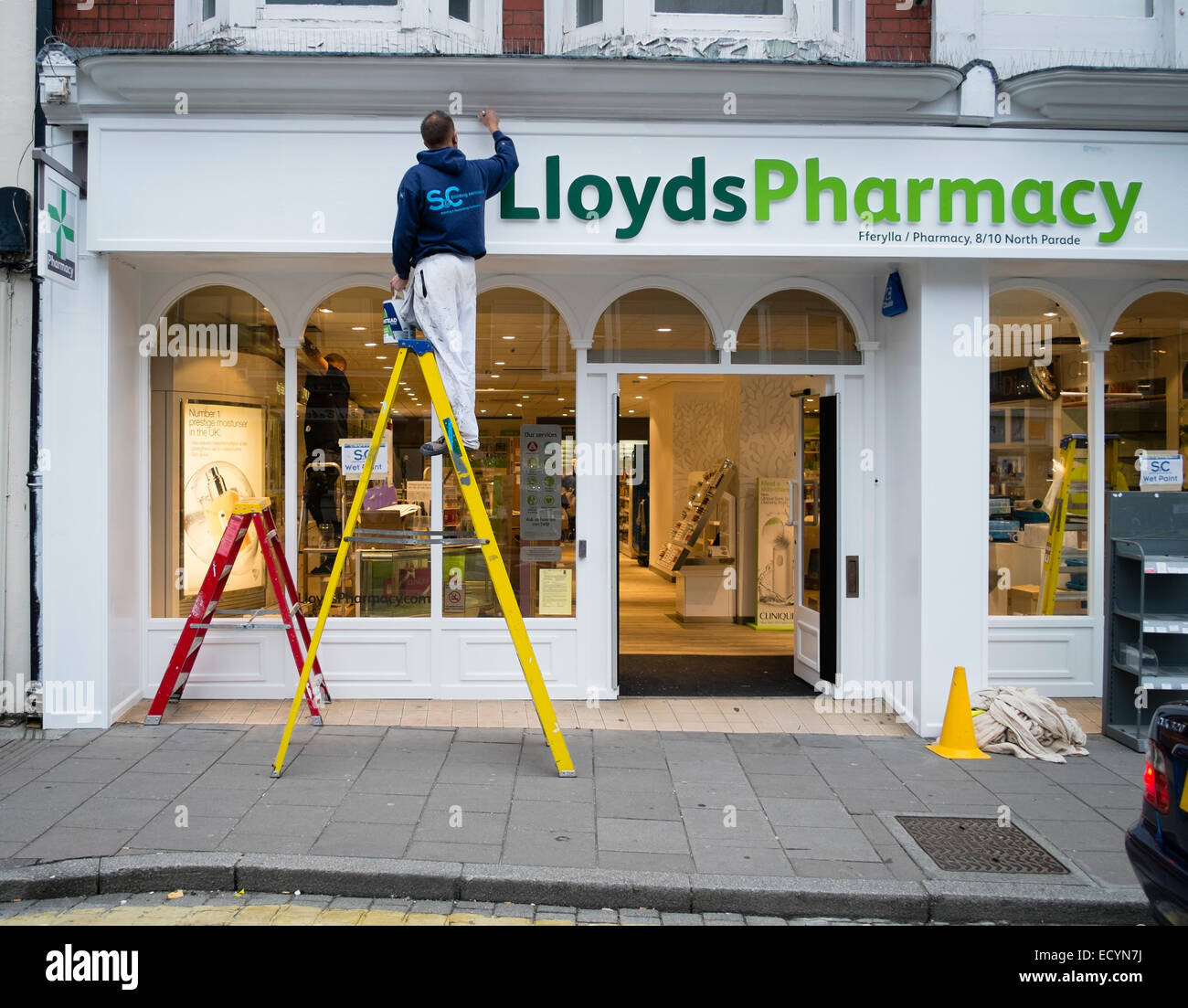 Vernice fresca: Un pittore in piedi su una scala verniciatura della parte esterna di una succursale della farmacia Lloyds Chemists shop store, REGNO UNITO Foto Stock