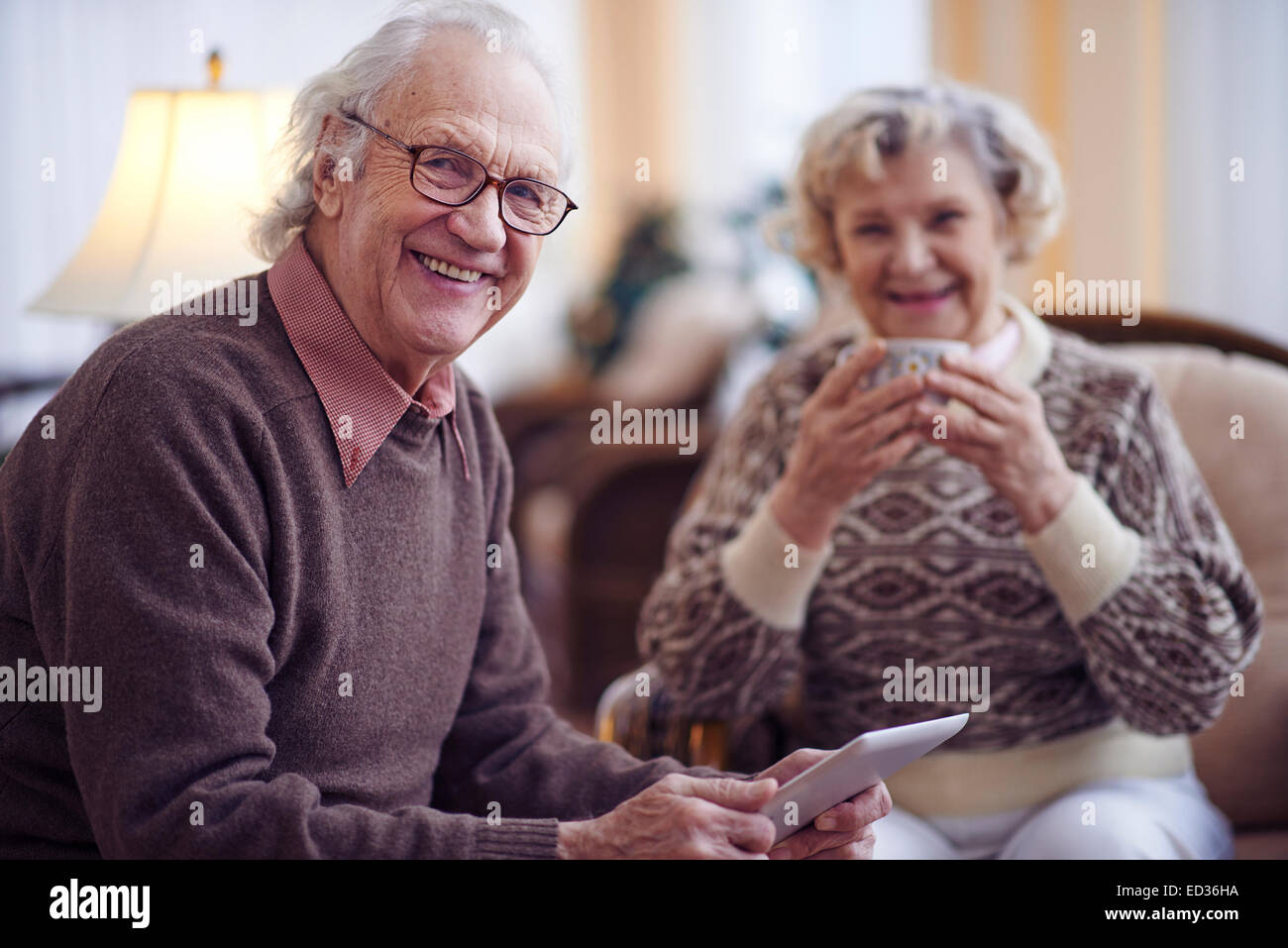 Uomo anziano con touchpad guardando la fotocamera con la moglie in background Foto Stock