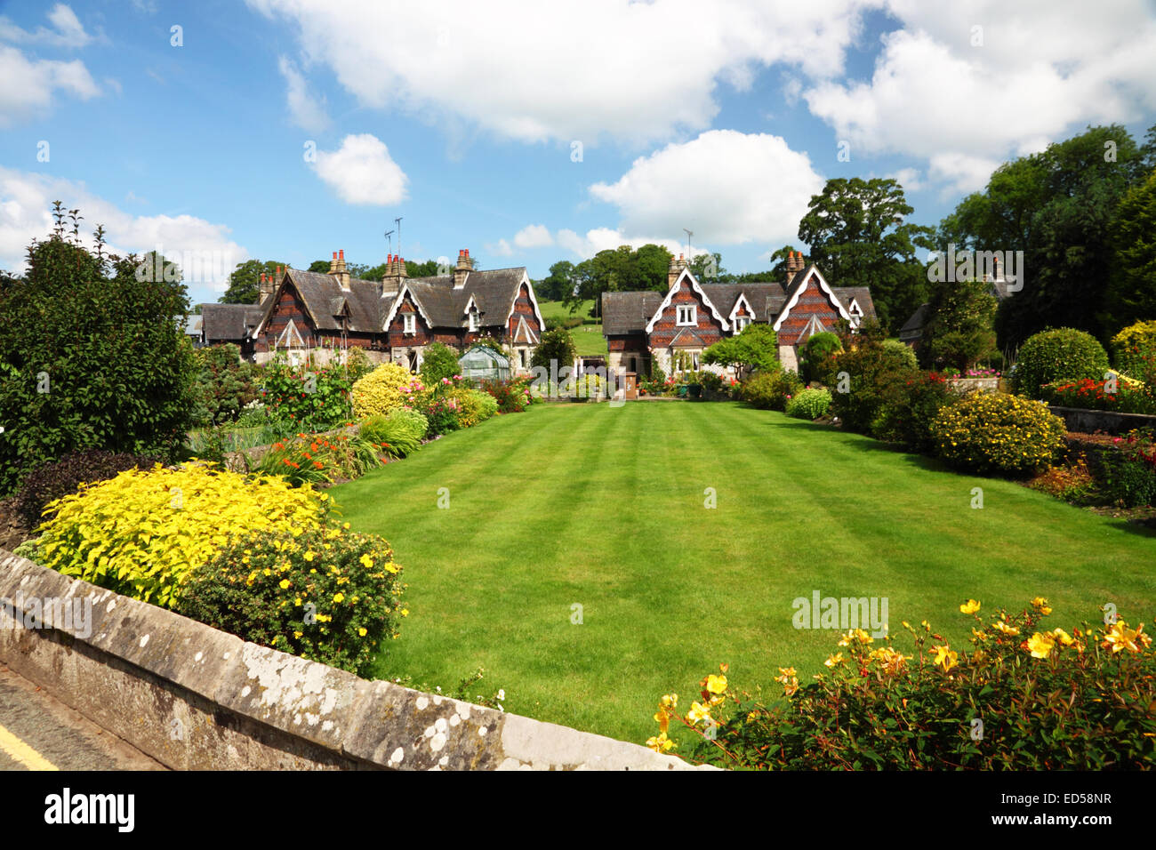 Un cottage Inglese giardino con un ampio prato, fiori di colore giallo e Swiss Cottage in stile sotto un cielo blu con nuvole. Foto Stock