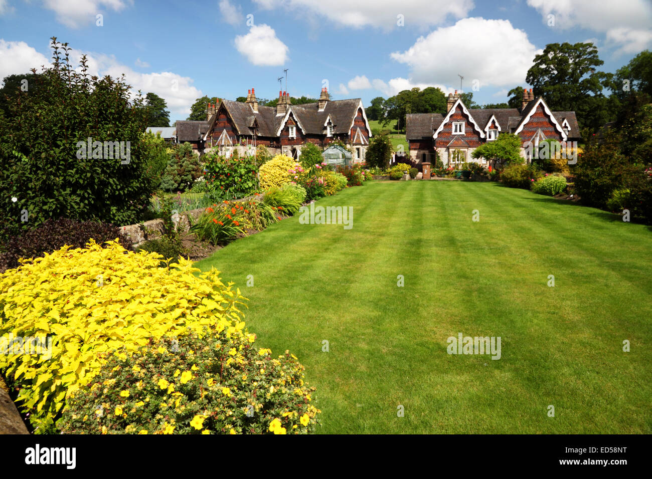 Un cottage Inglese giardino con un ampio prato, fiori di colore giallo e Swiss Cottage in stile sotto un cielo blu con nuvole. Foto Stock