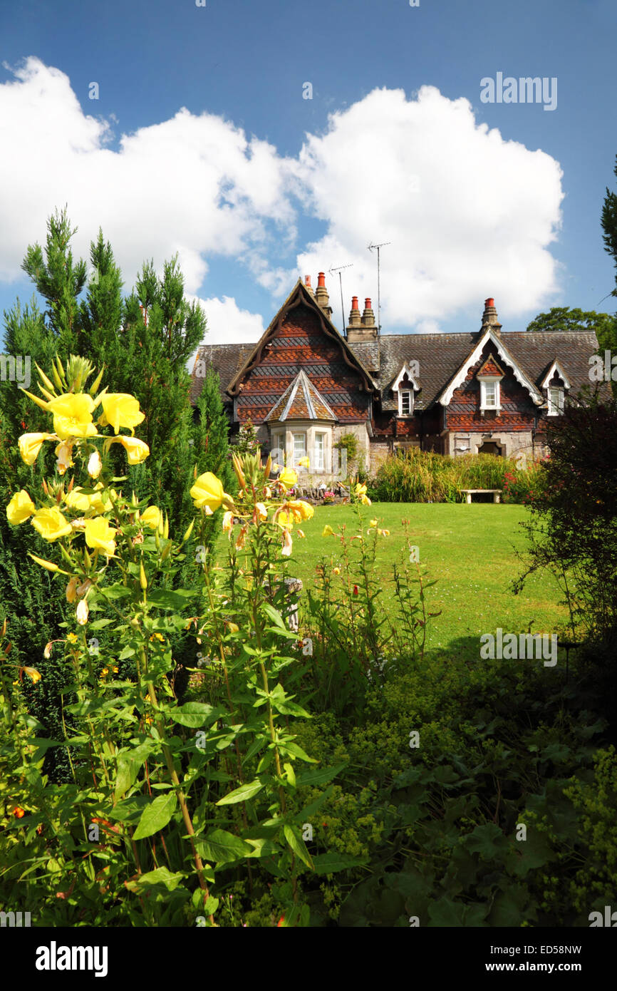 Un cottage Inglese giardino con prato, fiori di colore giallo e Swiss Cottage in stile sotto un cielo blu con nuvole. Foto Stock