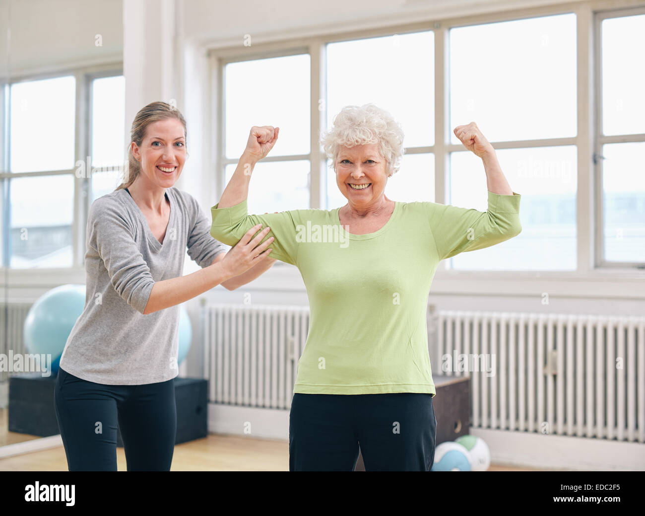 Ritratto di montare la vecchia donna flettendo le braccia e mostrando i suoi muscoli con personal trainer alla Palestra Foto Stock