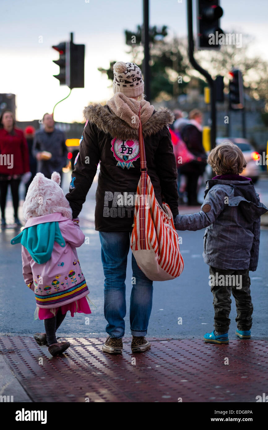 Vista posteriore di una giovane madre attraversare una strada trafficata tenendo le mani dei suoi due figli nella città di Cardiff, la capitale del Galles, Regno Unito Foto Stock