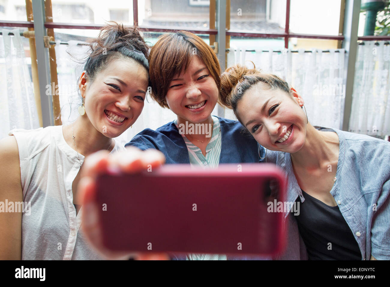 Tre donne alla ricerca di un telefono cellulare, prendendo un selfie, seduta all'interno. Foto Stock