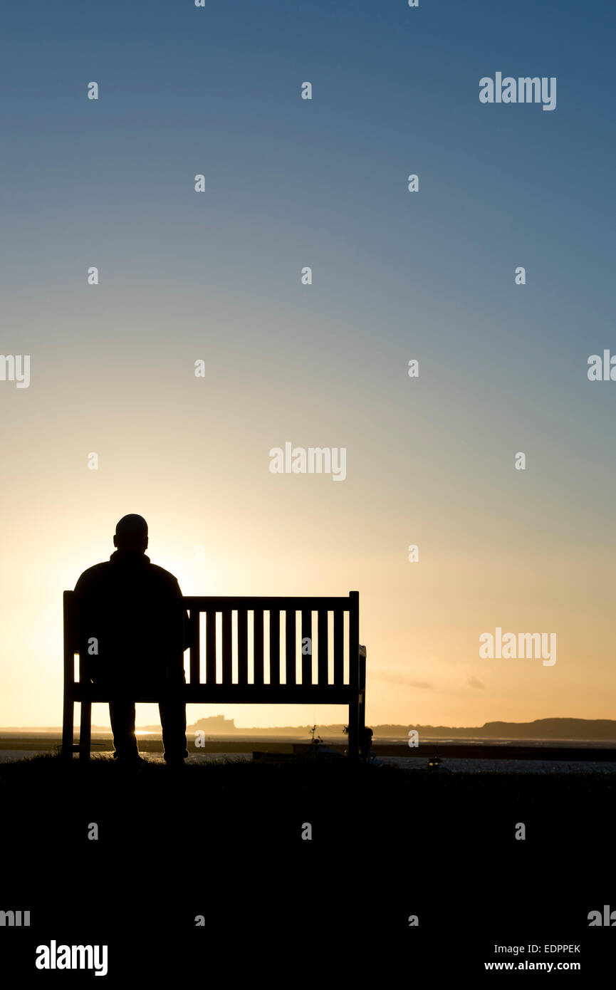 Sunrise silhouette di un uomo seduto su una panchina con il castello di Bamburgh nella distanza. A Isola Santa, Northumberland, Inghilterra. Foto Stock