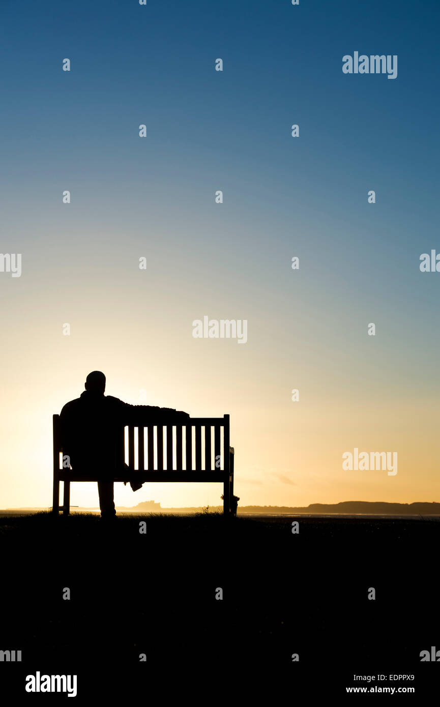 Sunrise silhouette di un uomo seduto su una panchina con il castello di Bamburgh nella distanza. A Isola Santa, Northumberland, Inghilterra. Foto Stock