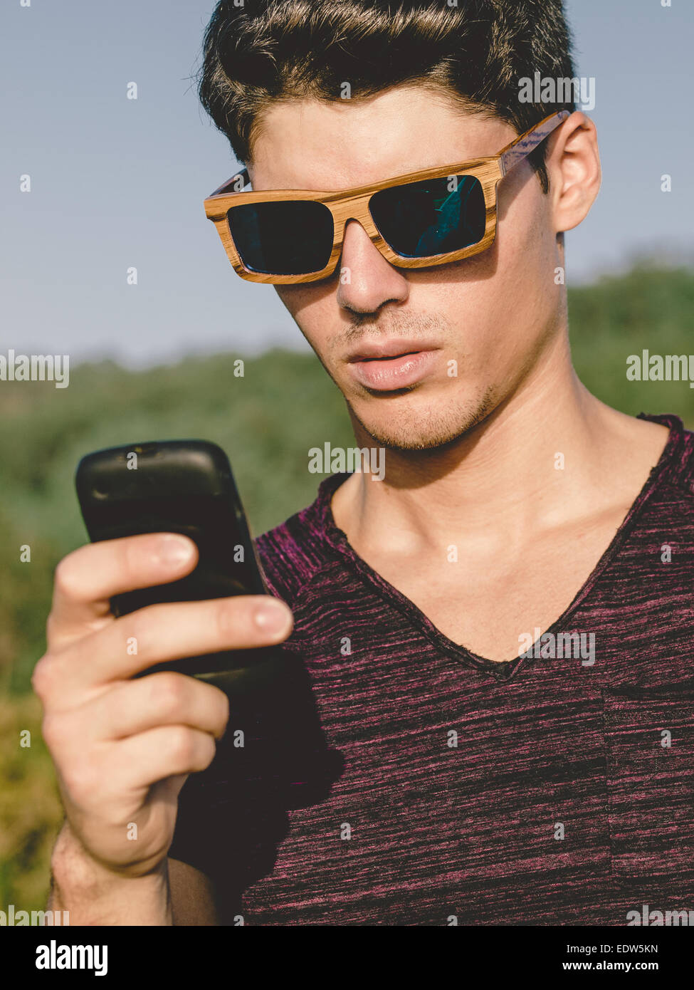 Modello uomo ritratto con occhiali da sole di legno all'aperto in una giornata di sole. L uomo è utilizzando uno smartphone Foto Stock