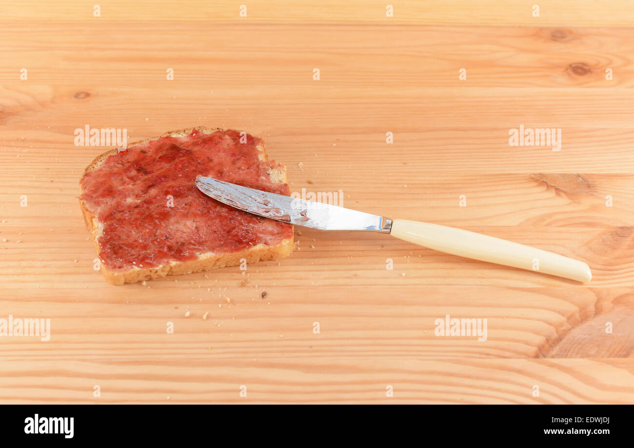 Diffusione di lampone o di confettura di fragole sul pane fresco con la lama di un coltello, appoggiato su un tavolo in legno Foto Stock