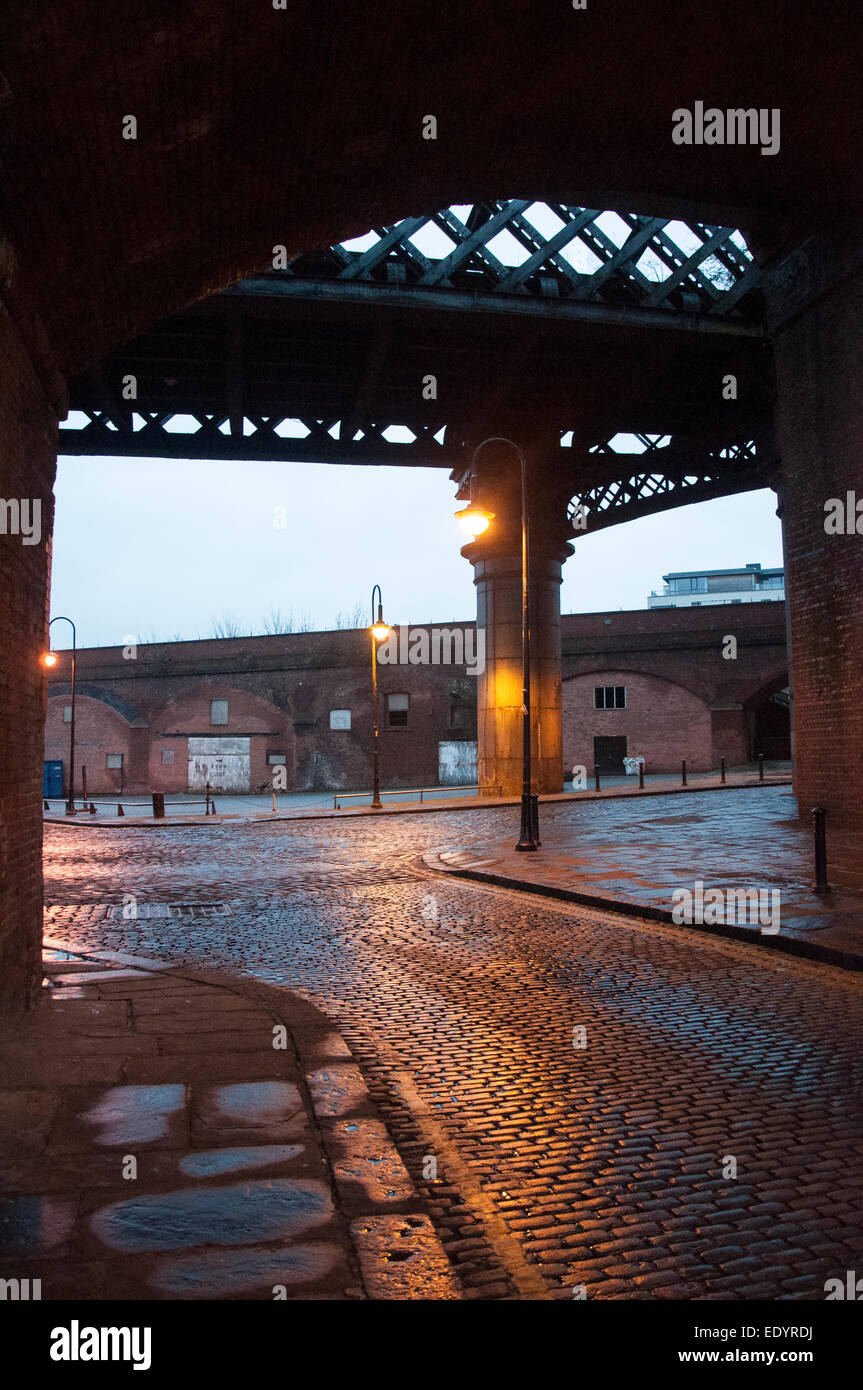 Vista serale attraverso un grande arco ferroviario vittoriano su ciottoli bagnati e magazzini di mattoni rossi a Castlefield di Manchester Foto Stock