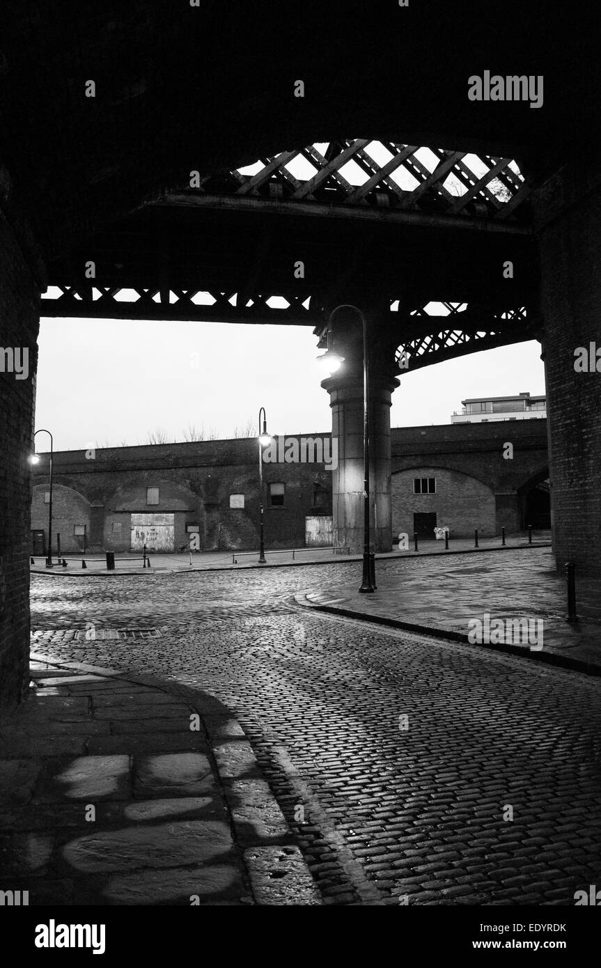 Vista serale in bianco e nero attraverso un arco ferroviario in ferro vittoriano su ciottoli bagnati ed edifici industriali in mattoni nel centro di Manchester. Foto Stock