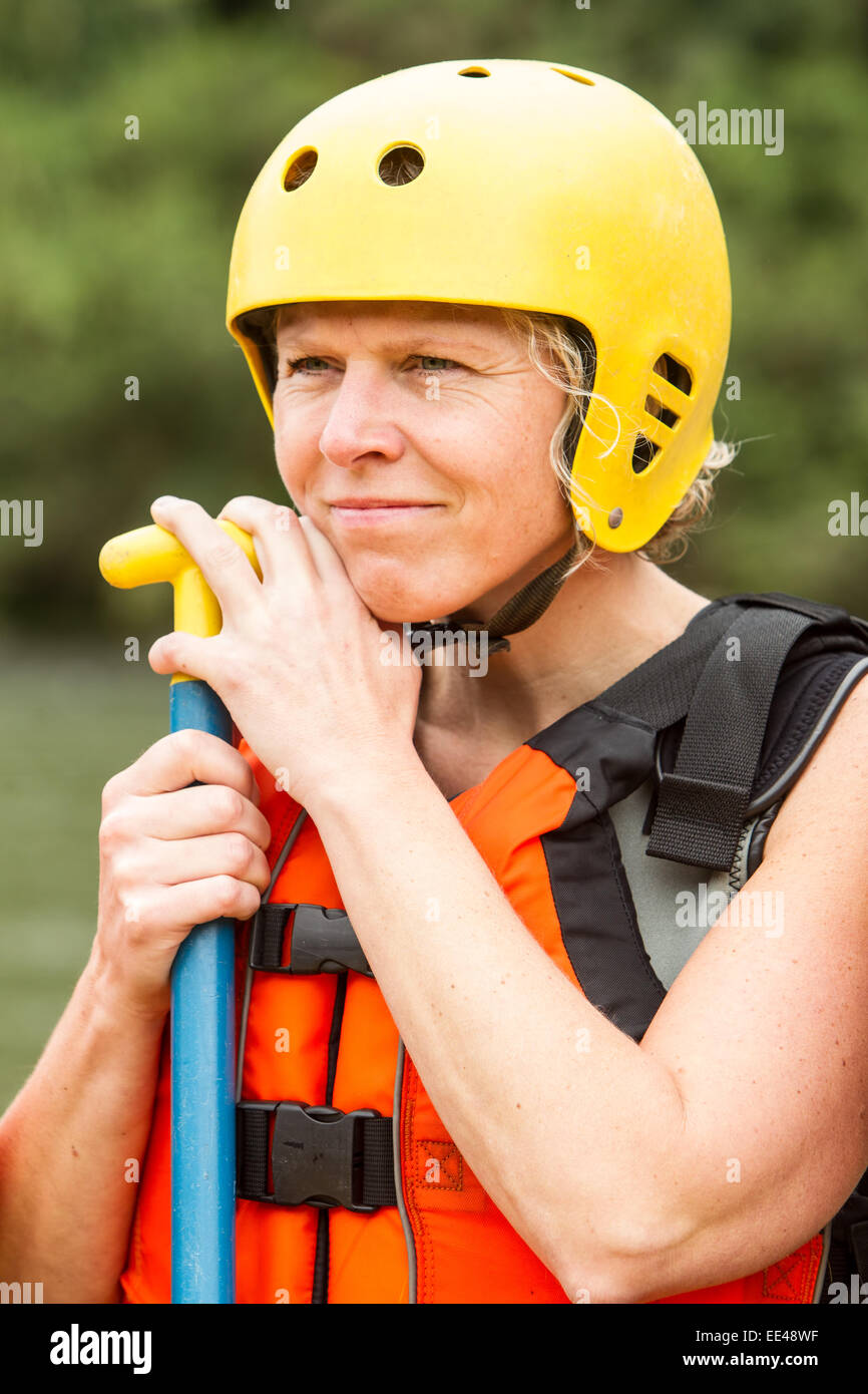 Una donna che indossa un casco di sicurezza giallo brillante e un giubbotto salvagente arancione regge una pagaia blu e gialla. Guarda leggermente di lato con un sorriso calmo e sottile. Le sue mani stanno afferrando il Foto Stock