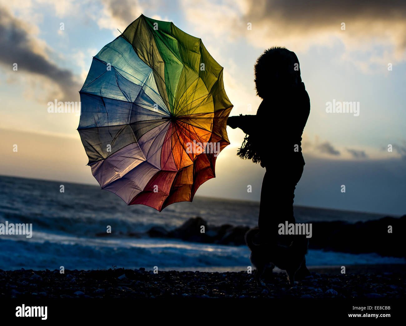 Una donna lotte con un ombrello in condizioni di vento forte come il sole tramonta sul mare. Foto Stock
