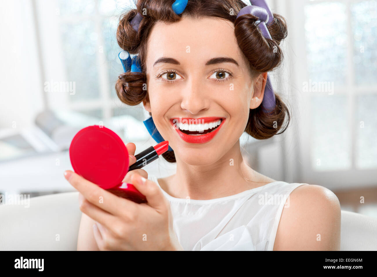 Donna sorridente con arricciacapelli utilizzando il rossetto bianco su sfondo domestico Foto Stock