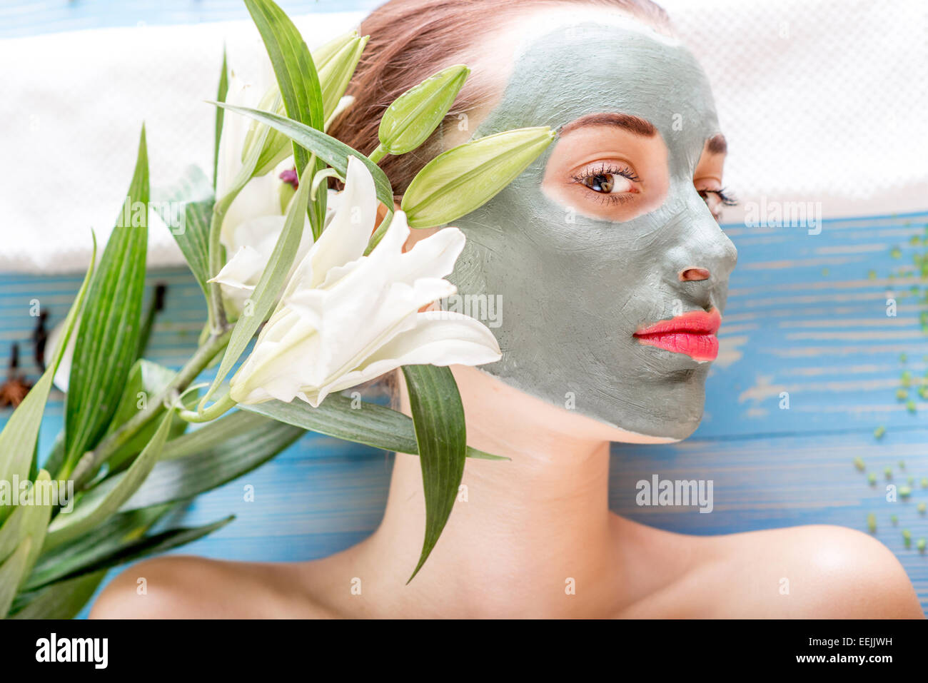 Giovane donna con centro termale viso maschera sul suo viso giacente sul tavolo blu con fiori, candele e sale marino in beaty salon Foto Stock