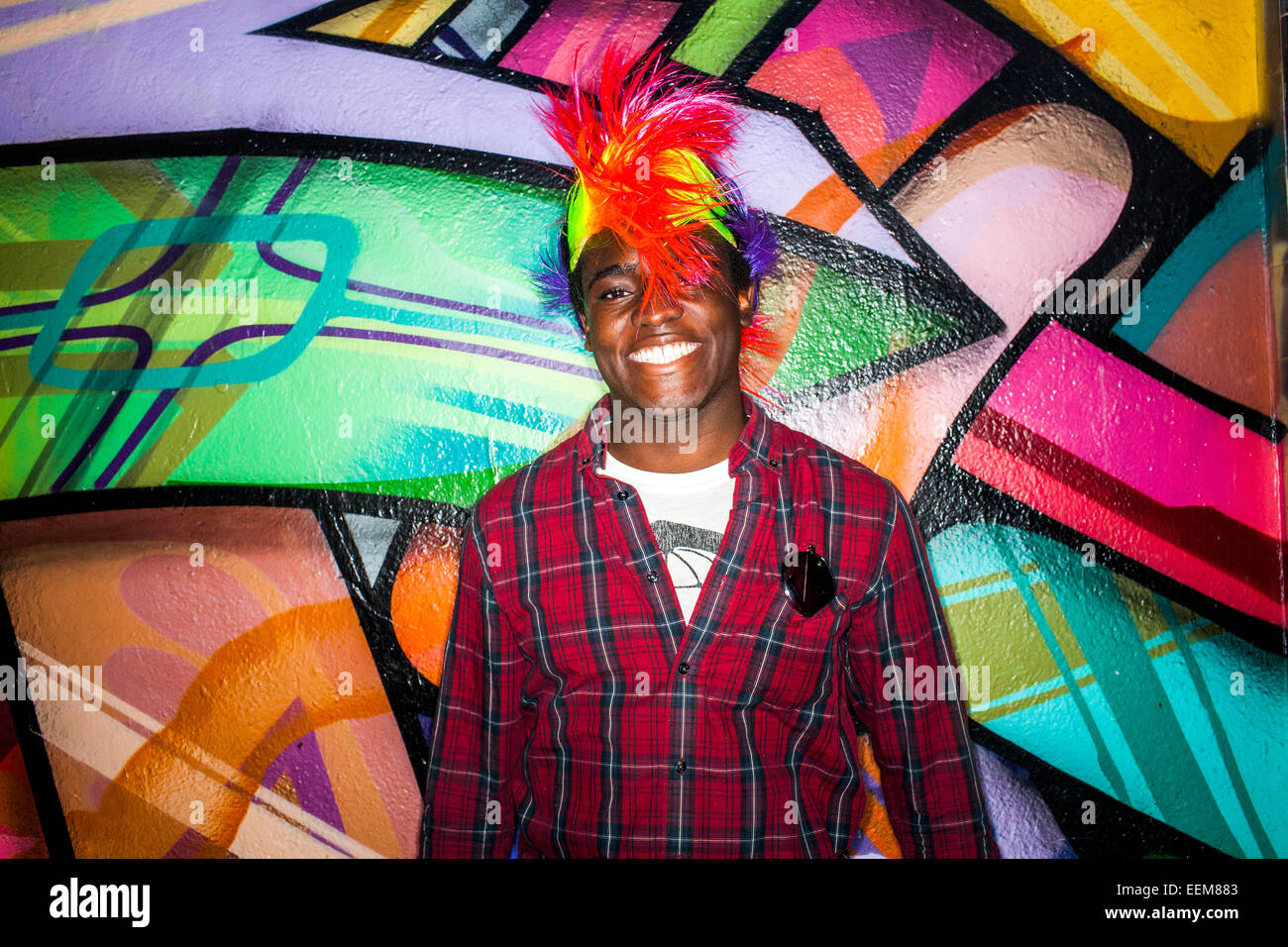 Uomo in nero parrucca colorata sorridendo vicino al muro di graffiti Foto Stock