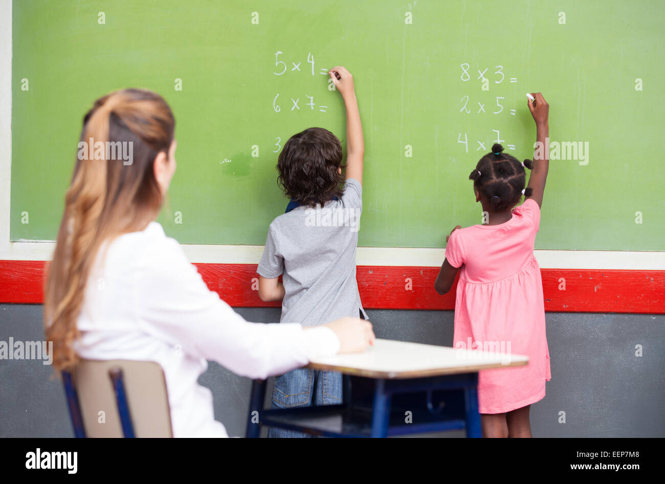 L apprendimento della matematica a scuola elementare. Multi etnico agli studenti di scrivere sulla lavagna con insegnante osservando. Foto Stock