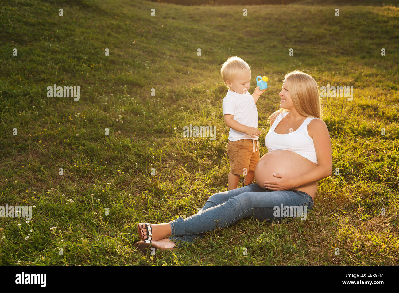 Carino piccolo ragazzo abbeveraggio di sua madre incinta pancia in estate la natura. Il grande fratello vuole la sua sorella a crescere più rapidamente. Festa della mamma Foto Stock