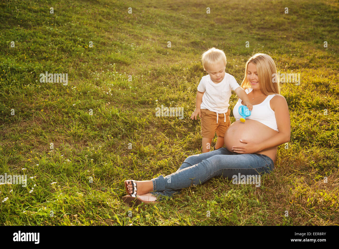 Carino piccolo ragazzo abbeveraggio di sua madre incinta pancia in estate la natura. Il grande fratello vuole la sua sorella a crescere più rapidamente. Festa della mamma Foto Stock