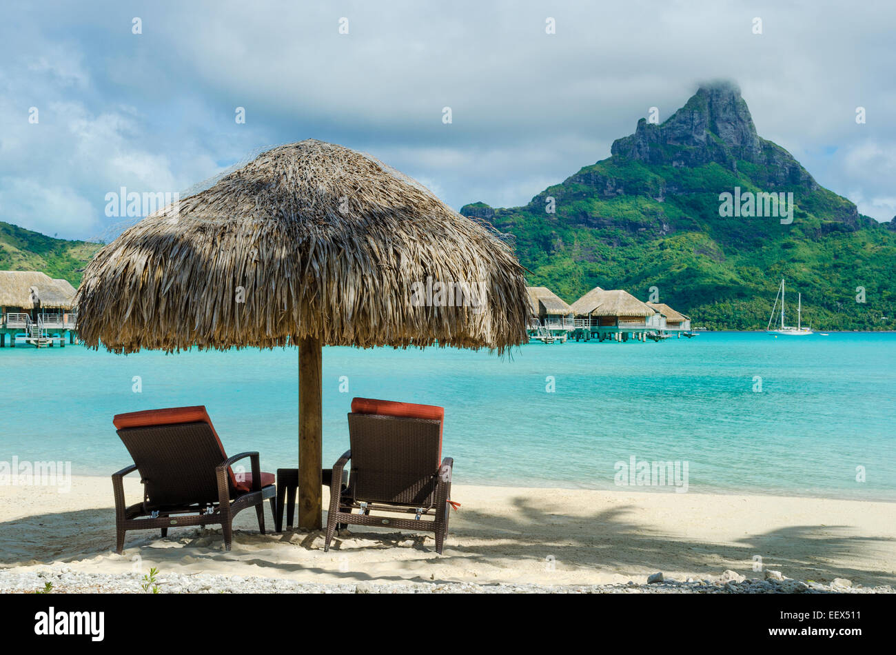 Due lettino sdraio sotto un ombrellone di paglia su una spiaggia di sabbia bianca con una vista sulla laguna e l'isola di Bora Bora. Foto Stock