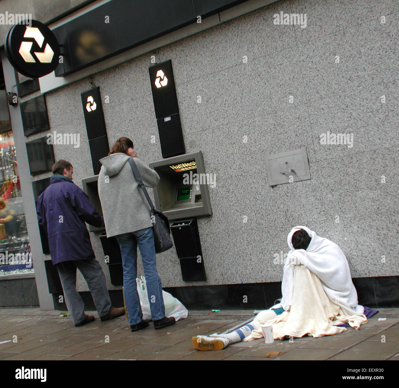 Divario ricco-povero e disuguaglianza sociale illustrato da un bancomat NatWest a Tottenham Court Road, Londra, con un disabile senza tetto coperto di coperte seduto nelle vicinanze Foto Stock