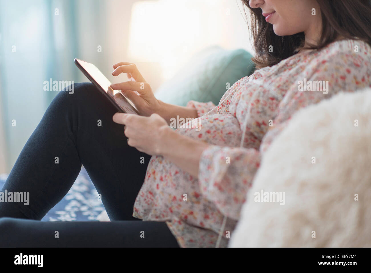 Donna incinta seduta sul letto tramite tablet Foto Stock